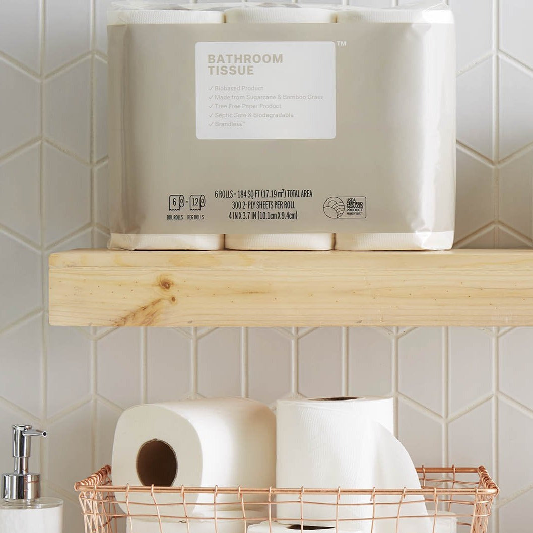 Lifestyle, six pack of bathroom tissue sites on a bathroom overjohn shelf above a copper wire basket and a hand soap dispenser. Bathroom Tissue: biobased product, made from sugarcane and bamboo, tree free paper product, spetic safe, brandless. Contains six 300-sheet rolls.