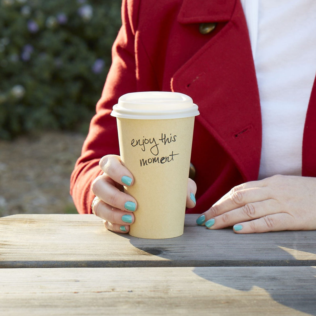 Lifestyle photo of a woman holding a cup of coffee while seated in a park with the inspirational message of &#39;enjoy this moment&#39; written ono the side of the cup.