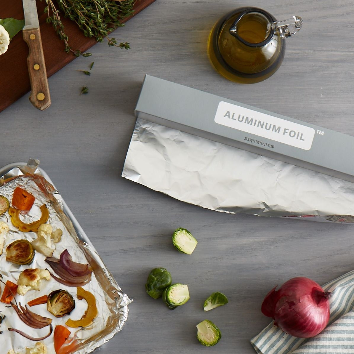 Lifestyle photo, aluminum foil on a kitchen counter showing a baking tray lined with foil and showing nicely roasted sliced brussel sprouts, onion quarters, and chunks of tomato.