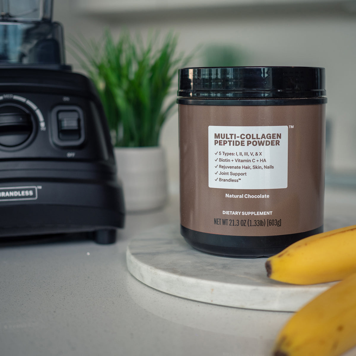 Lifestyle photo, tub of multi-collagen peptide powder sits on a kitchen counter next to a brandless Pro-Blender and some ripe bananas.