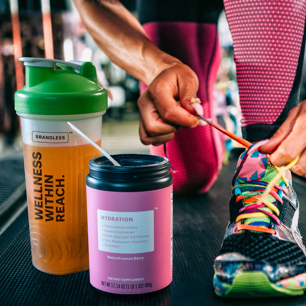 Lifestyle photo, runner in bright neon colors kneels on a gym floor to tie a shoelace next to a shaker bottle with lemon berry hydration mixed in it.