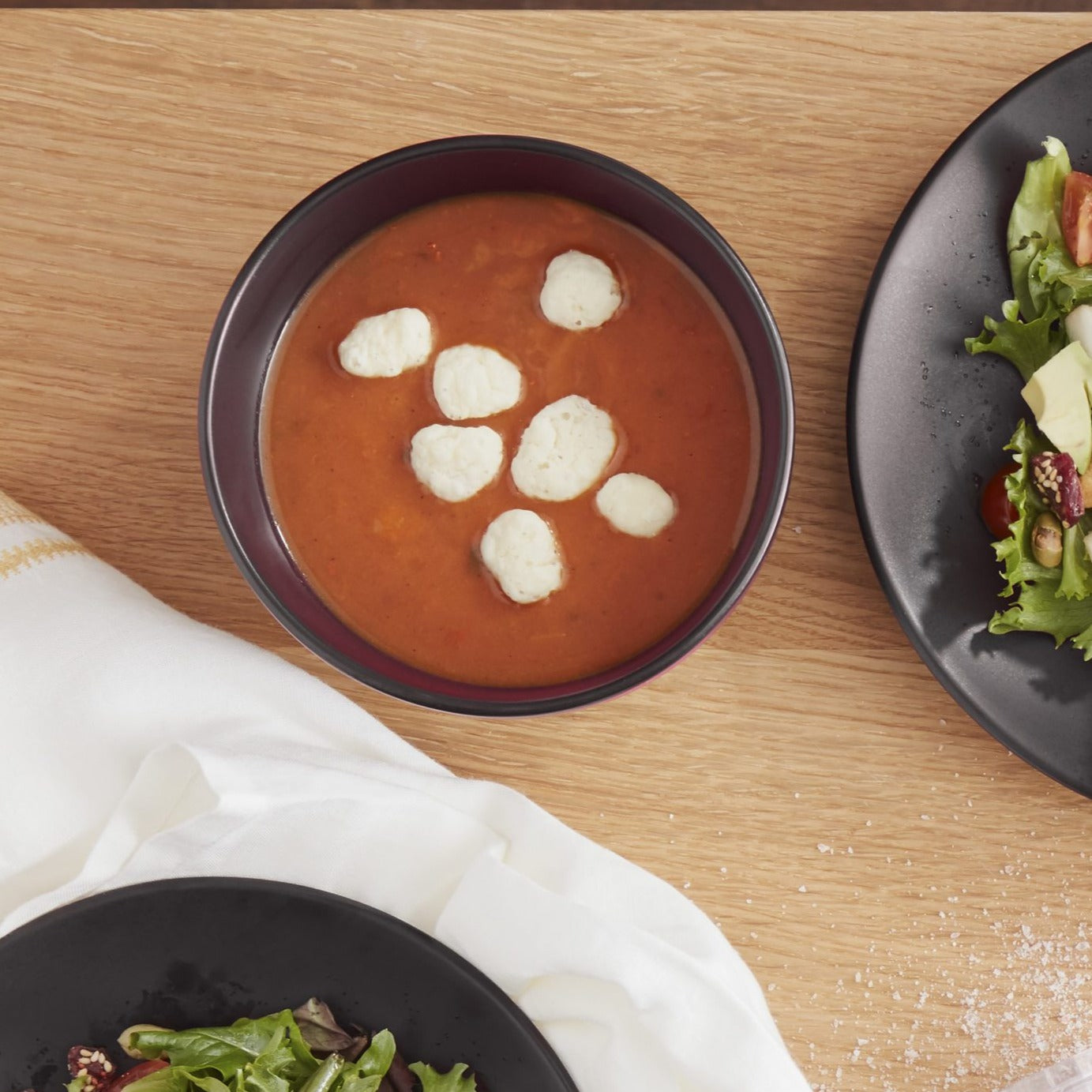 Lifestyle photo, black stoneware cereal bowl with parmasean cheese puffs topping a tomato bean soup sitting on a kitchen table next to salad plates with a cranberry sesasme salad on them.