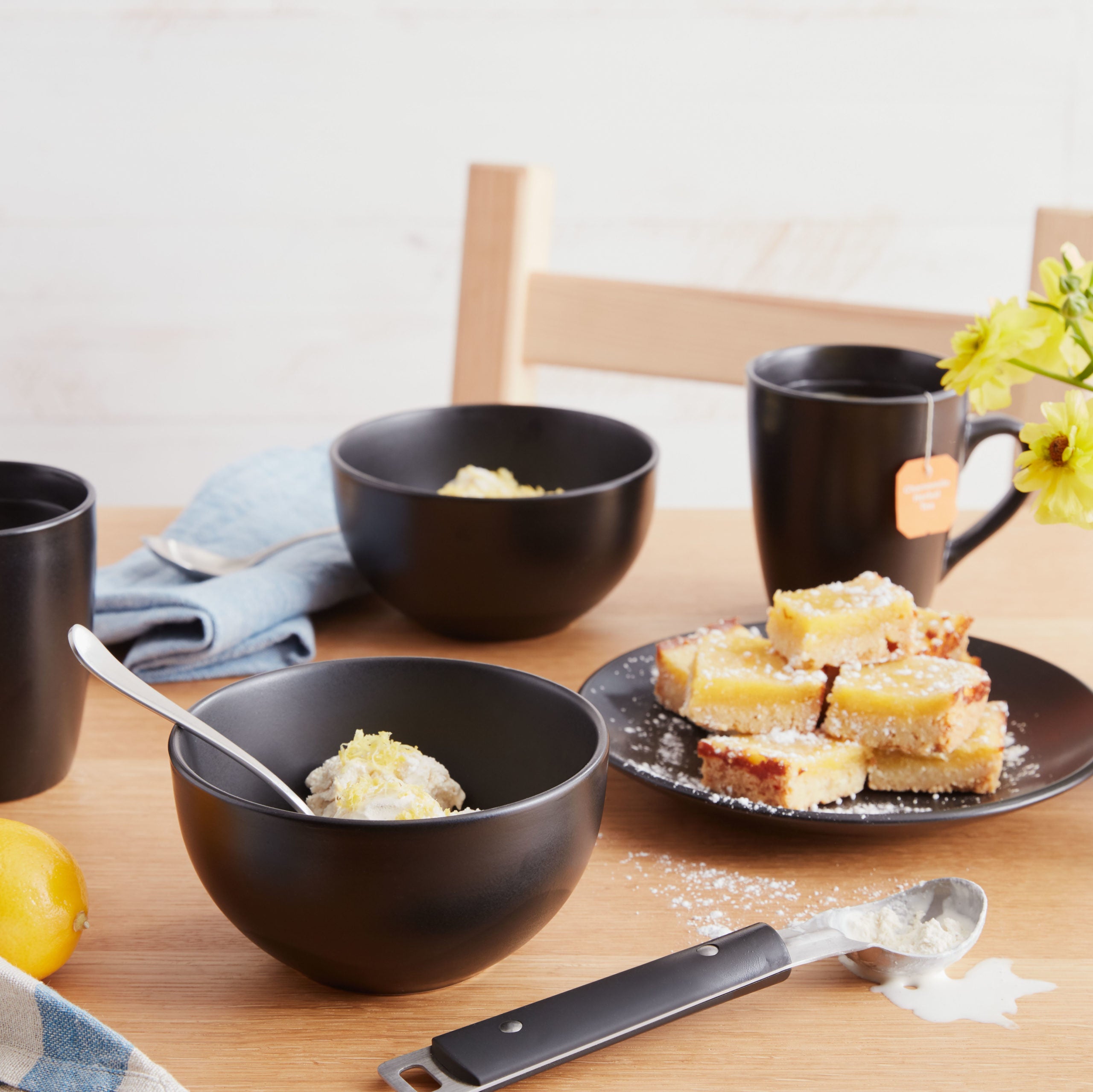 Lifestyle photo, ice cream spoon, black stoneware cereal bowl with ice cream topped with lemon zest. Black stoneware salad plate with a pile of lemon bars topped with powdered sugar. Two black stoneware mugs with tea bags steeping.