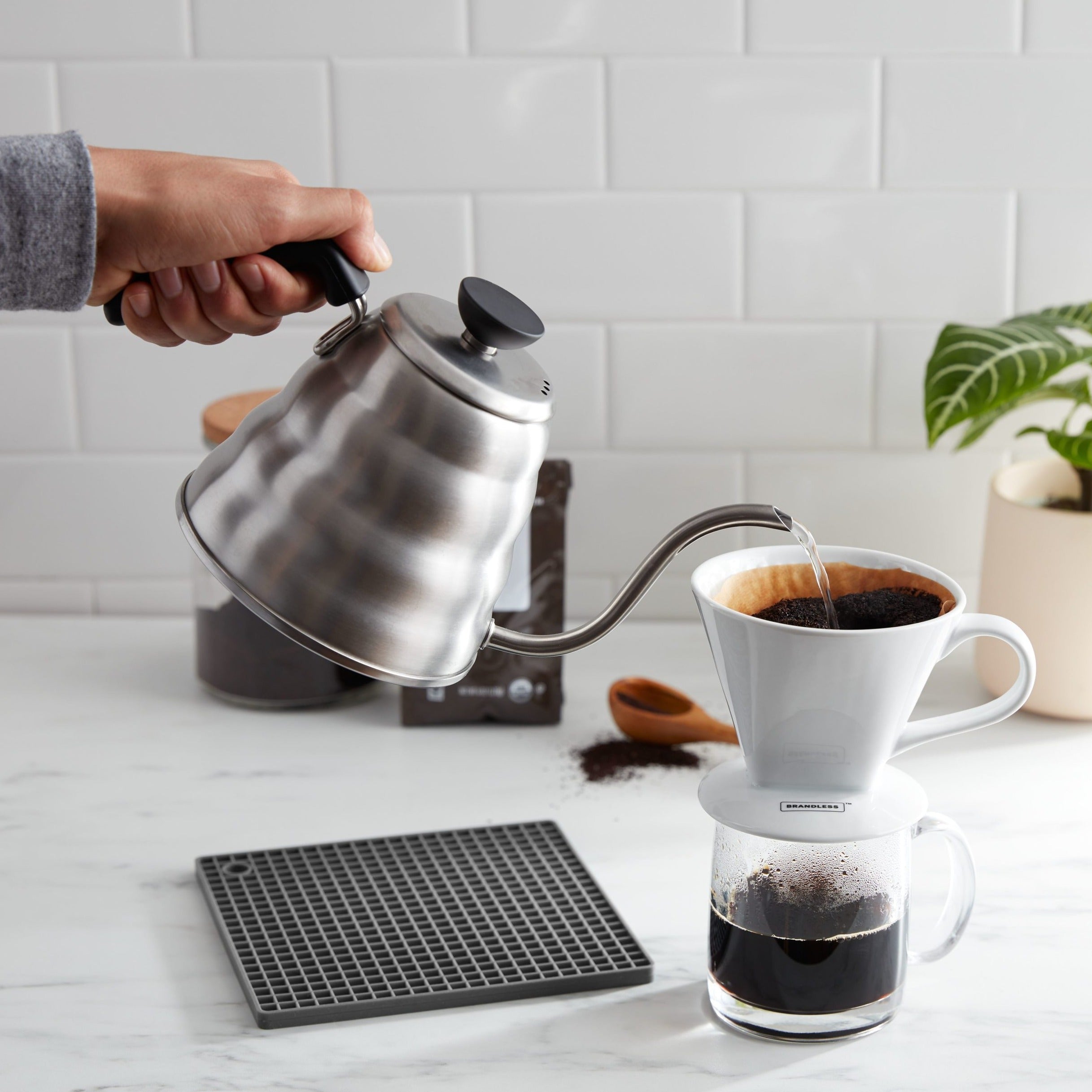 Lifestyle photo, hand shown pouring hot water from a tea kettle over fresh grounds in a paper filter in the coffee cone to create a hot, perfectly-mixed brew.