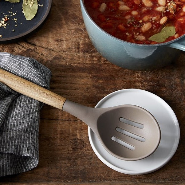 Lifestyle photo, acacia wood handled silicone slotted serving spoon resting on a spoon rest next to a pot of chili.