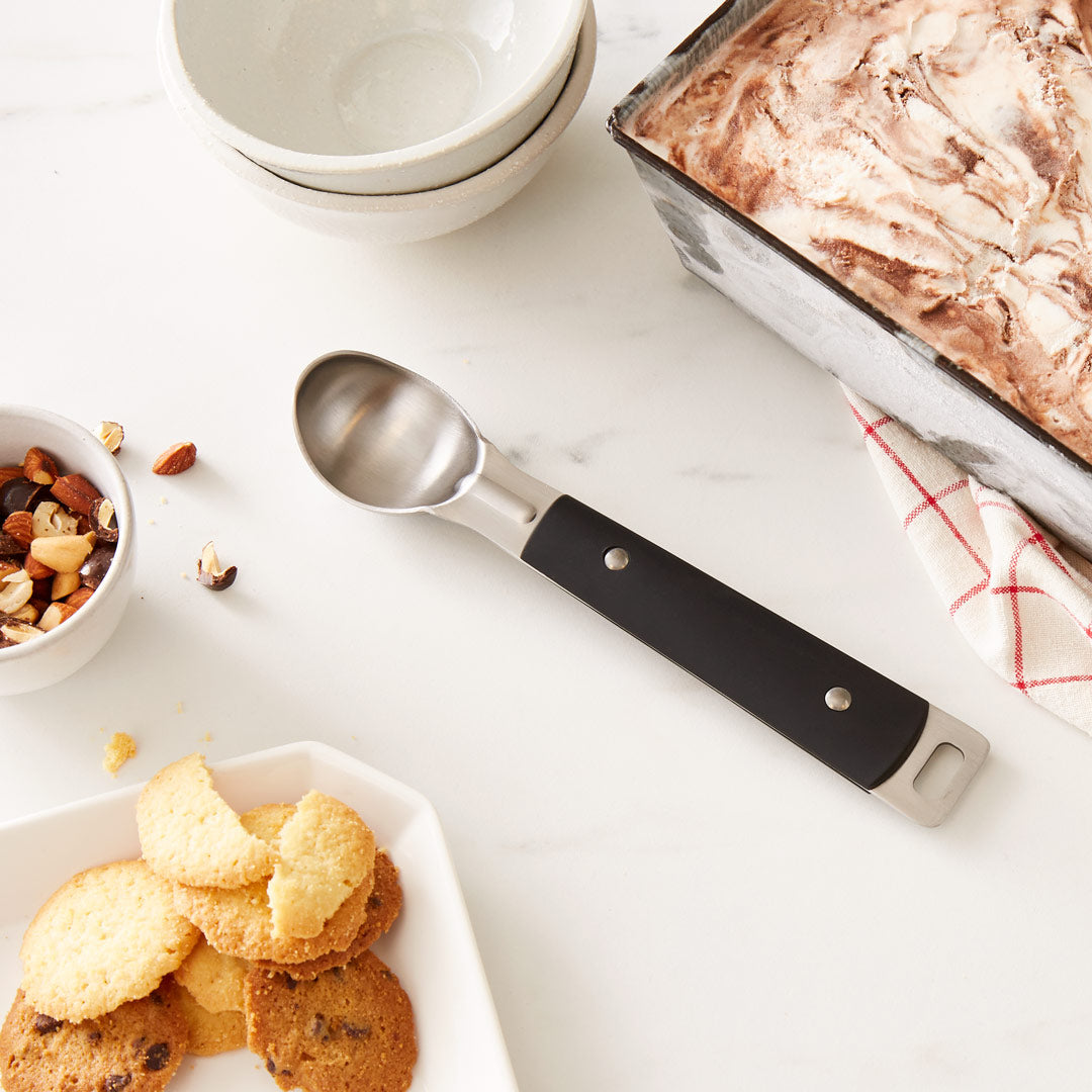 Lifestyle photo, ice cream scoop on a kitchen counternext to a dish of homemade ice cream, and some nuts in a small bowl ready to springkle on top.
