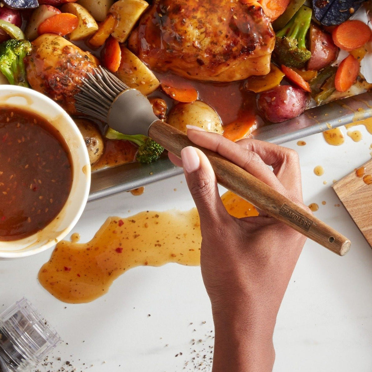 Lifestyle photo, womans&#39;s hands using silicone basting brush spreading a sweet chili marinade over a baking tray of chicken separates and chopped veggies.
