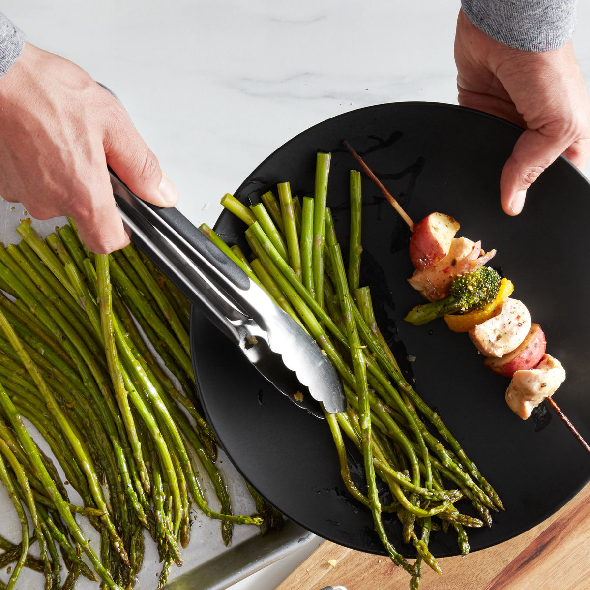 Lifestyle photo, Man&#39;s hands using 9 inch stainless steel tongs transferring some roasted asparagus spears from a baking tray to a dinner plate.