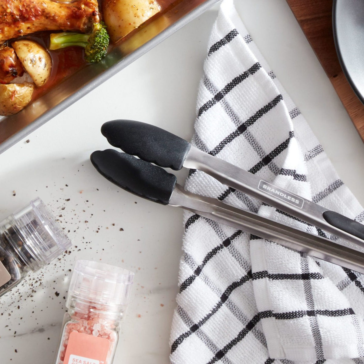 Lifestyle photo, silicone baking tongs in closed and locked position sitting on a kitchen counter next to a baking tray with chicken and vegetables on it and a kitchen towel near it.