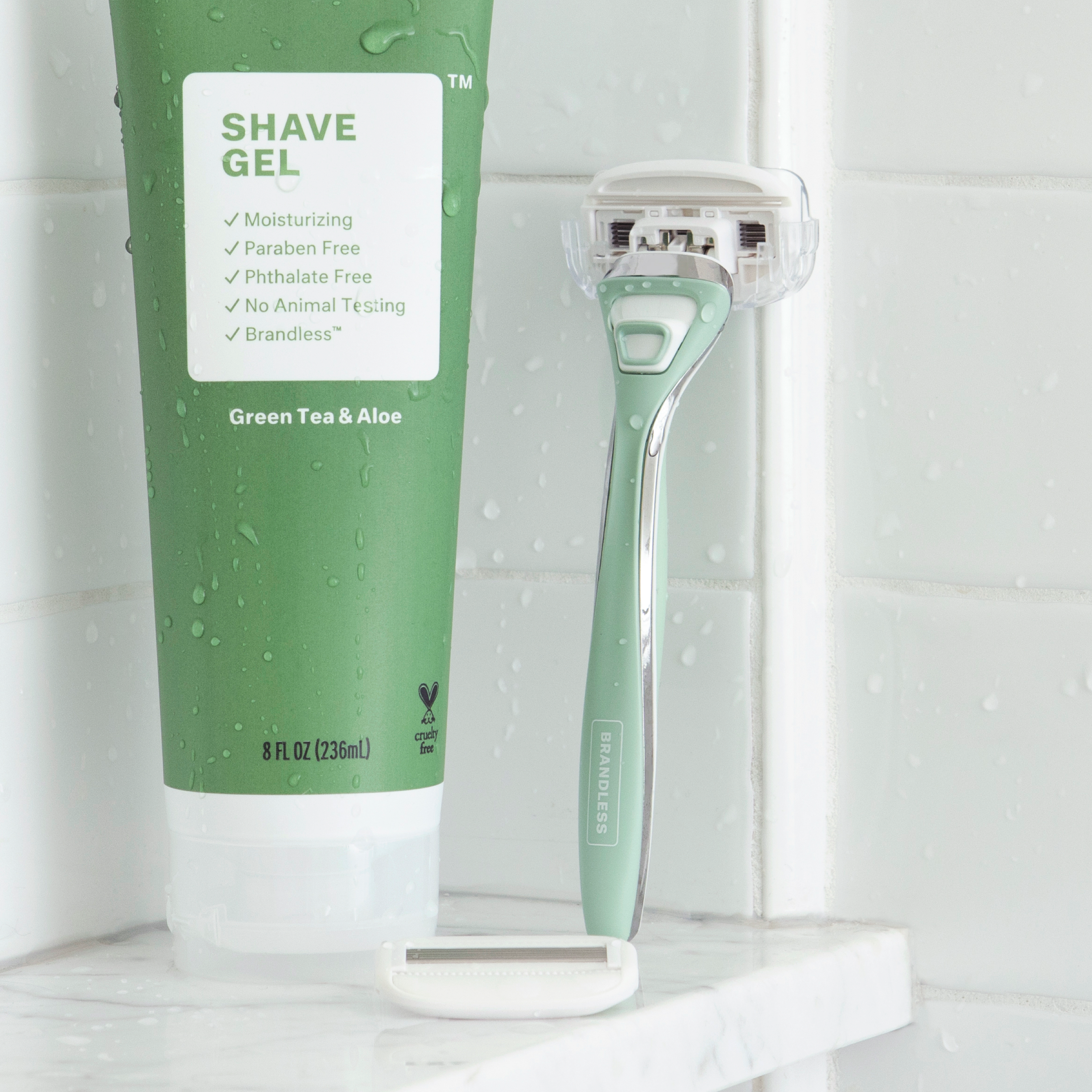 Lifestyle photo, razor for body stands up against the tile wall of a shower on a shelf next to a tube of brandless green tea and aloe shave gel. Water droplets abound.