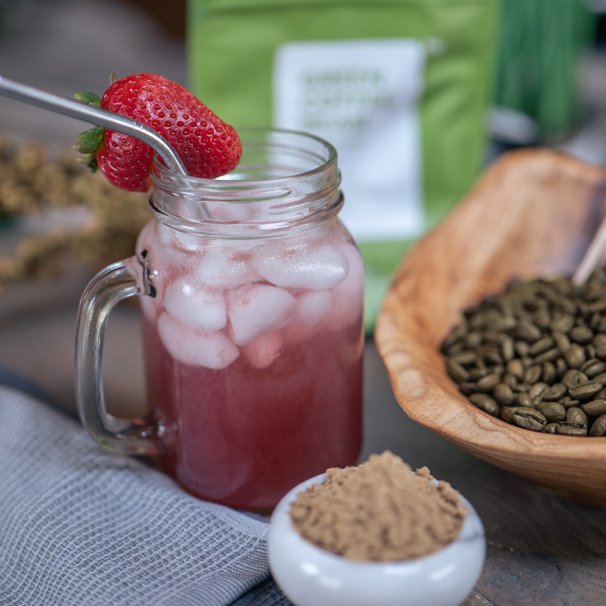 Lifestlye photo of a pink drink with ice and strawberries in a mason jar.  Green coffee beans held in a wood container.