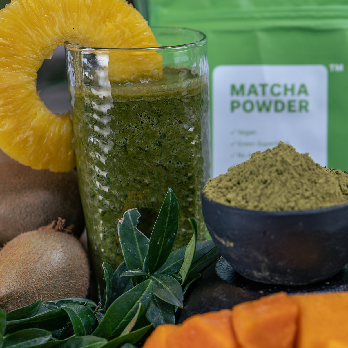 Lifestyle photo, rich green matcha smoothie in glass next to pineapple chunks, kiwi, matcha powder.