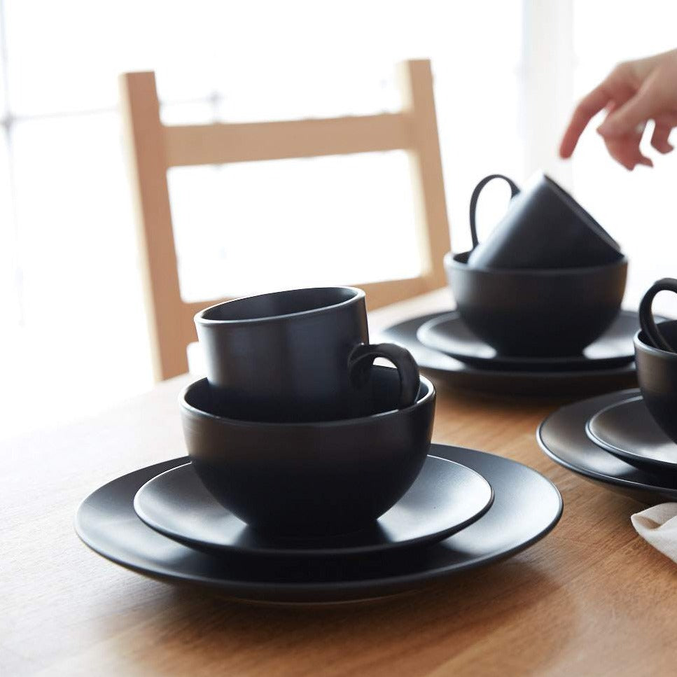 Lifestyle photo, stacked stoneware service sets of a dinner plate, salad plate, cereal bowl, and mug. Three piles of stoneware decorate a kitchen table and a hand reaches in to pick up a mug.