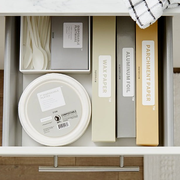 Lifestyle photo, showing Brandless's line of kitchen baking accessories: parchment paper, aluminum foil, wax paper, tree-free paper plates, and disosable flatware all stored neatly in a kitchen drawer.