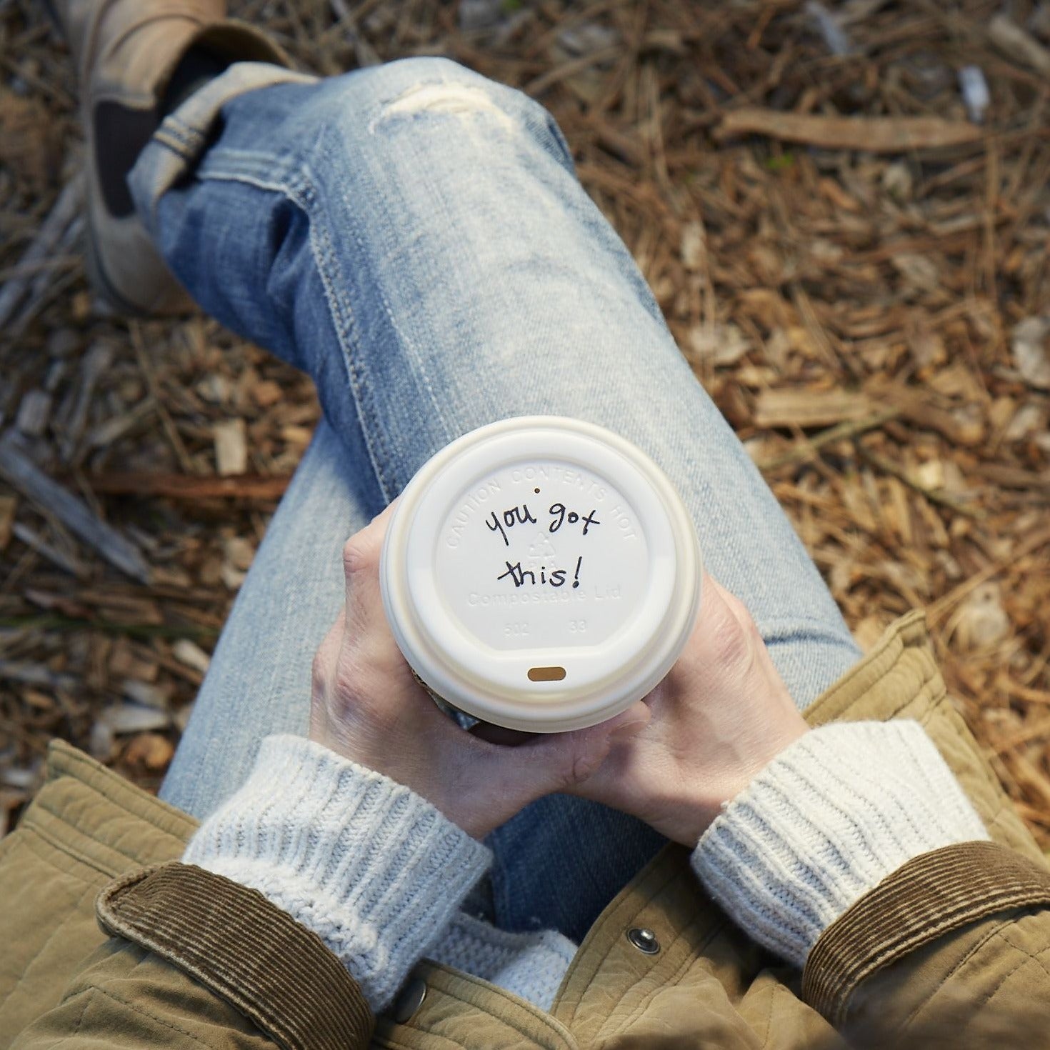 Lifestyle photo of a person holding a cup of coffee while seated in a park with the inspirational message of  'you got this!' written ono the lid.