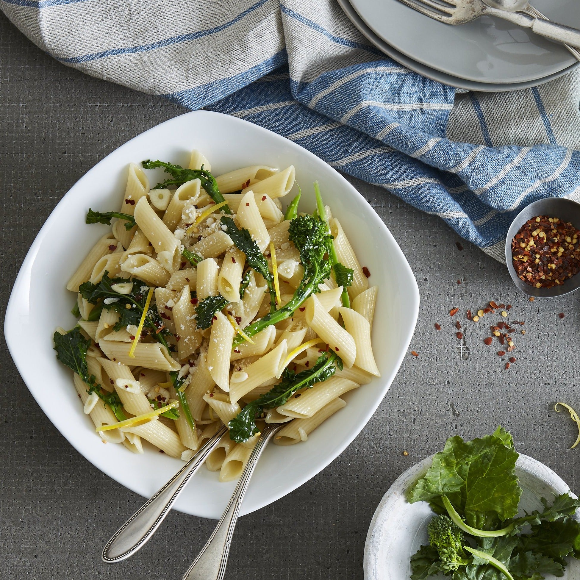 Lifestyle. Top view of a kitchen counter as a chef plates a meal.  Serving bowl is full of a fresh penne and kale pasta dish with parmasean and chilli flakes on top, ready to serve beautifully with a pair of silver serving forks.