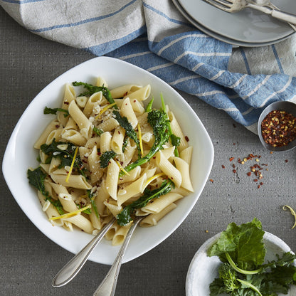 Lifestyle. Top view of a kitchen counter as a chef plates a meal.  Serving bowl is full of a fresh penne and kale pasta dish with parmasean and chilli flakes on top, ready to serve beautifully with a pair of silver serving forks.