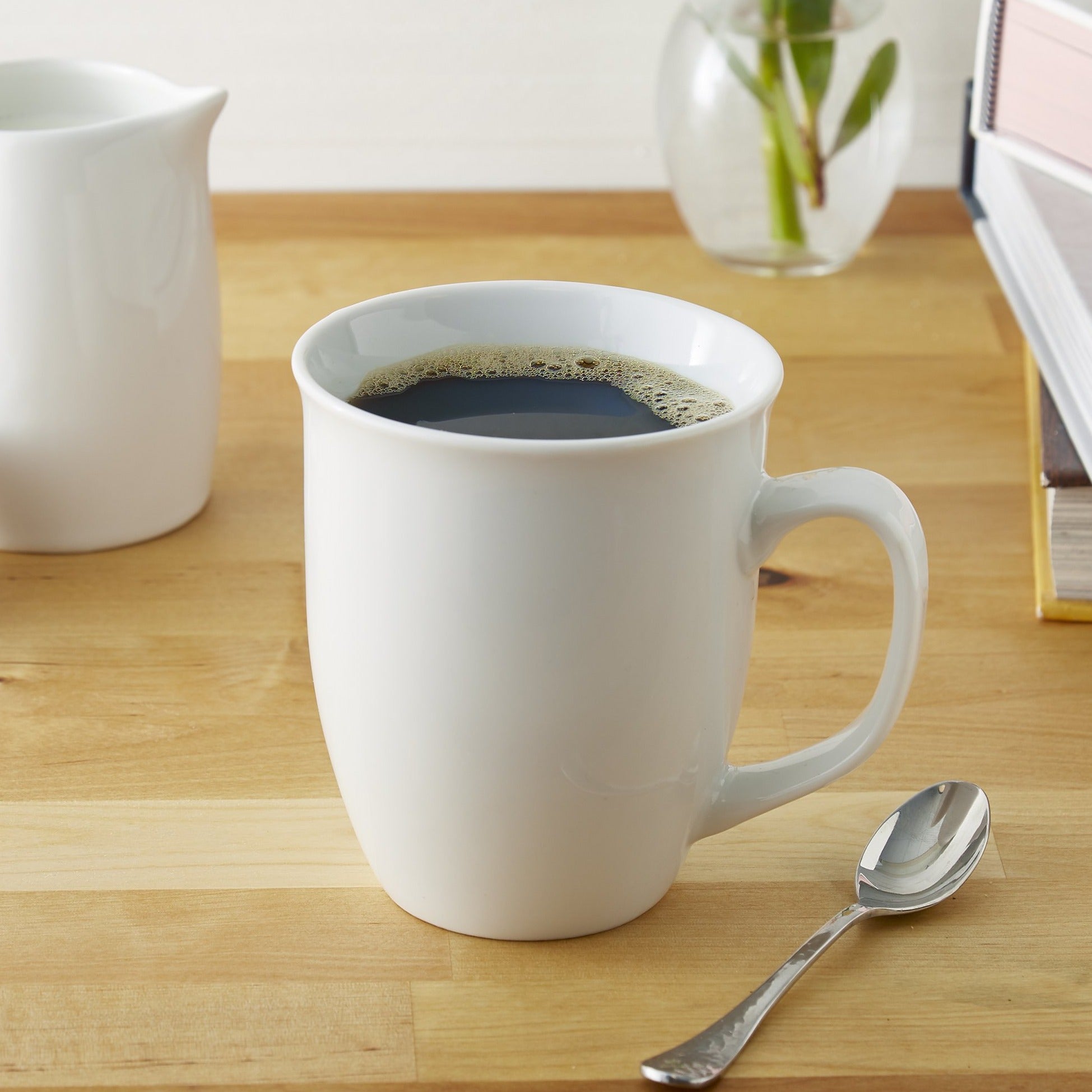Lifestyle photo, white porcelain coffee mug with a fresh cup of steaming coffeee in it sitting on a kitchen counter next to a spoon and some reading material.