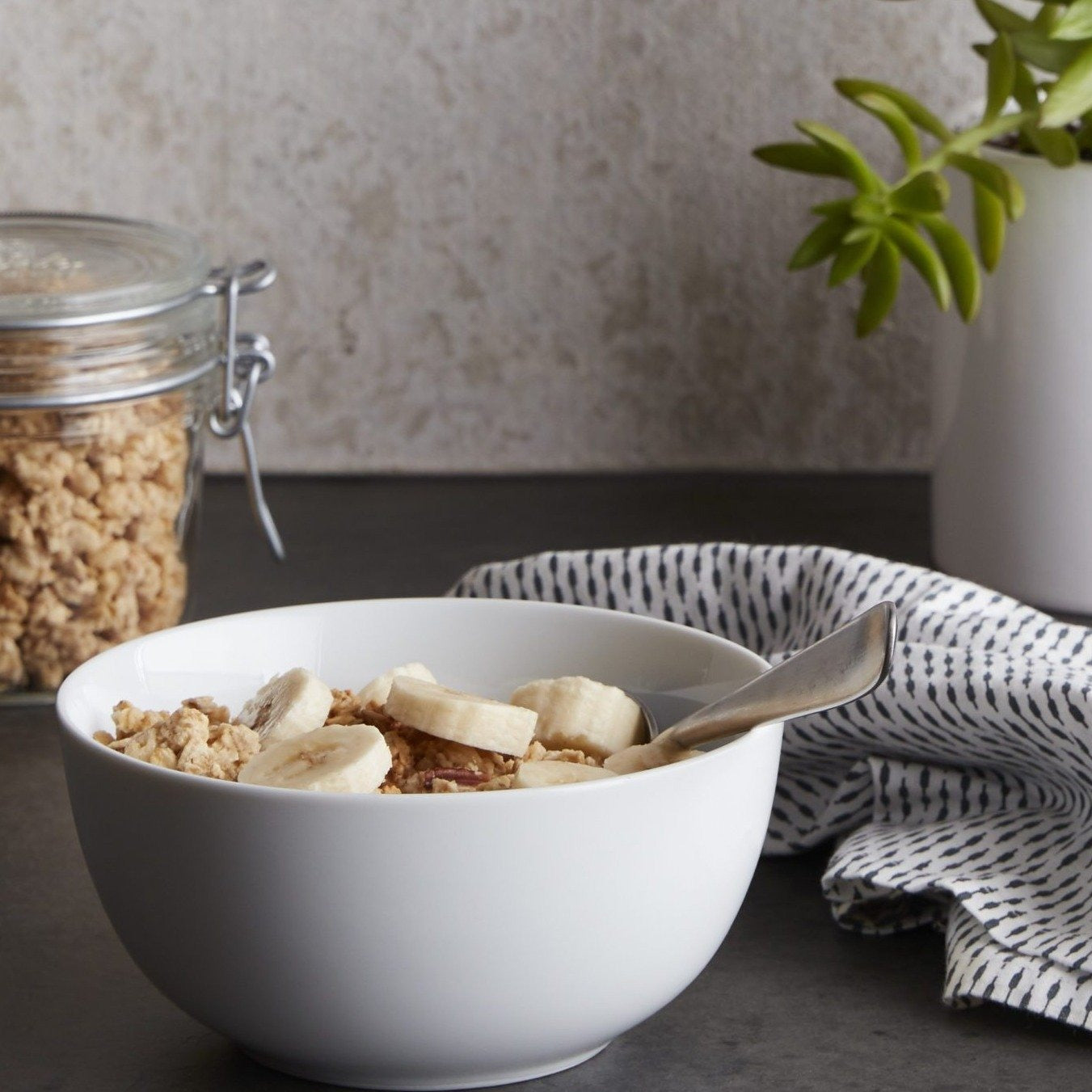 Lifestyle photo, cereal bowl with granola and fresh sliced banana sits on a kitchen counter next to a dish towel, plant, and jar full of granola.