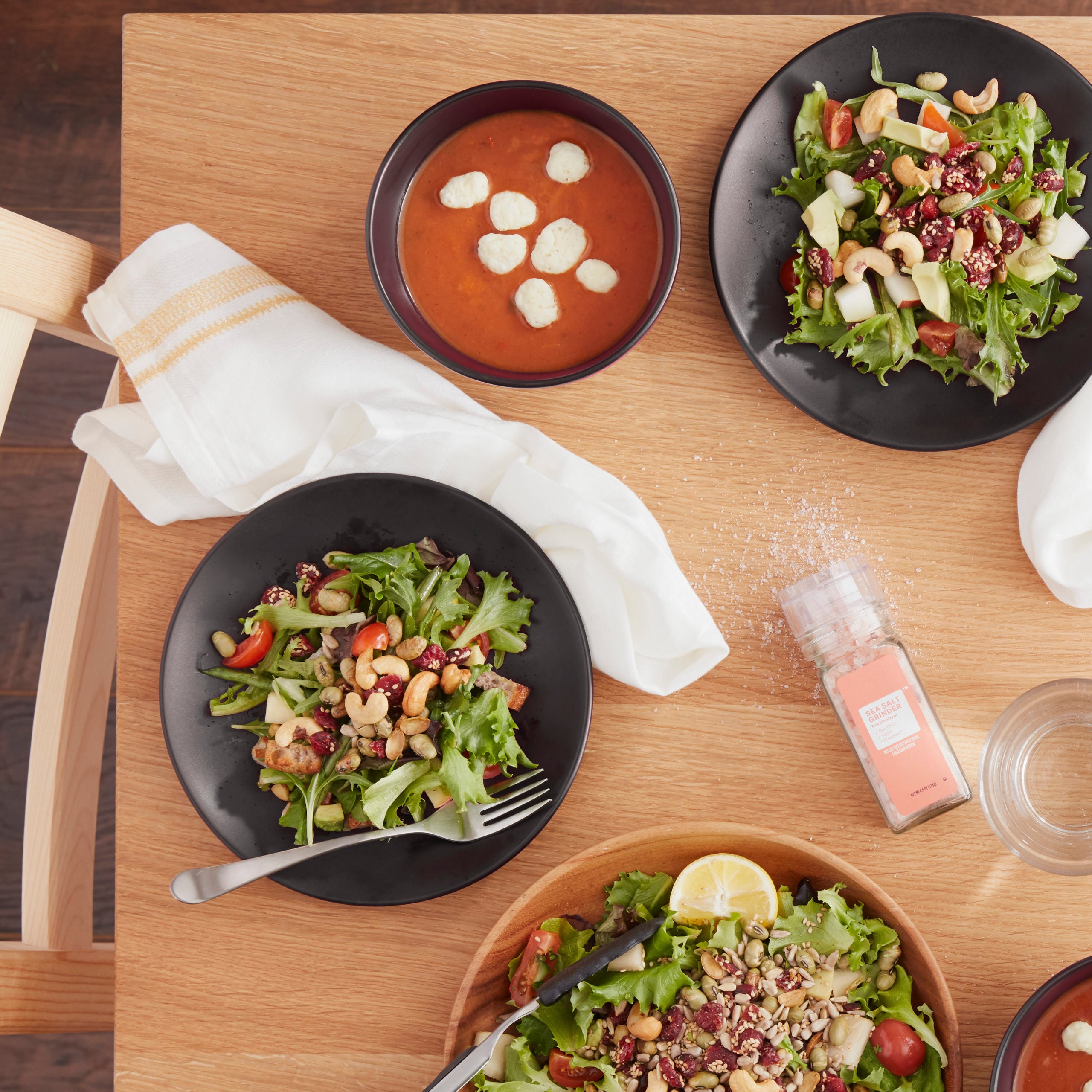Lifestyle photo, black stoneware cereal bowl with parmasean cheese puffs topping a tomato bean soup sitting on a kitchen table next to salad plates with a cranberry sesasme salad on them.