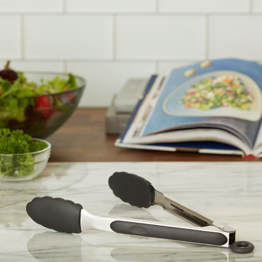 Lifestyle photo, 9 inch silicone baking tongs in their open position laying on a marble kitchen counter next to a bowl of sprigs about to be put on top of a tossed salad as decoration.