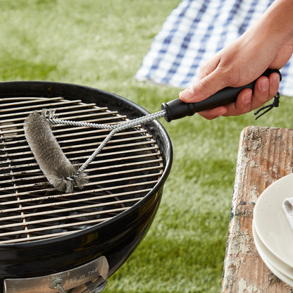 Lifestyle photo, backyard, hand holding a grill brush up next to a charcoal grill