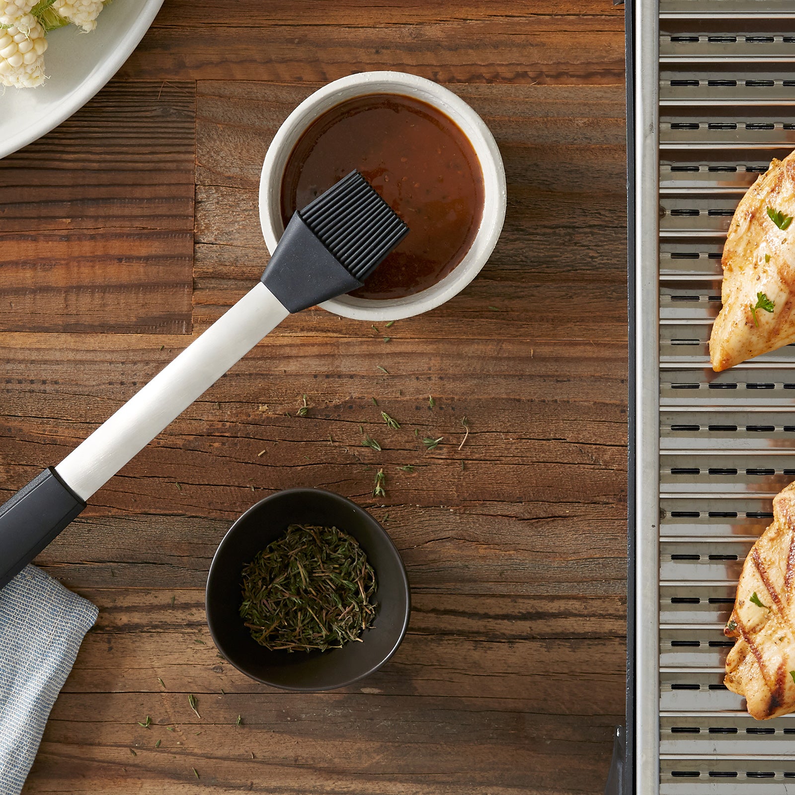 Lifestyle photo. Grill basting brush leaning on a small bowl with a savory red sauce, getting prepared to spread sauce on some freshly grilled items.
