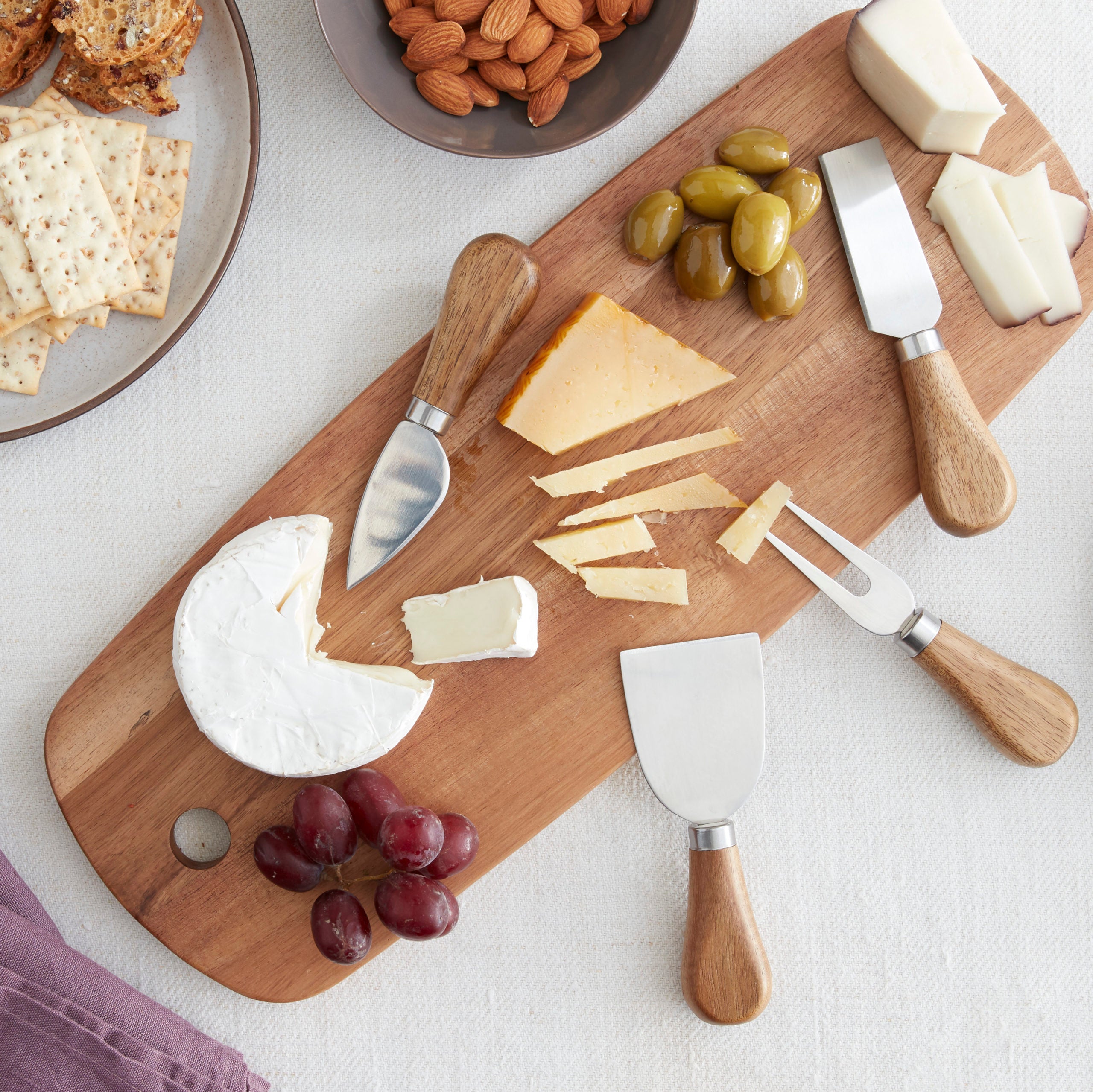 Lifestyle photo, acacia cheese board with tapas preparation ingredients including grapes, three types of cheese, and olives.