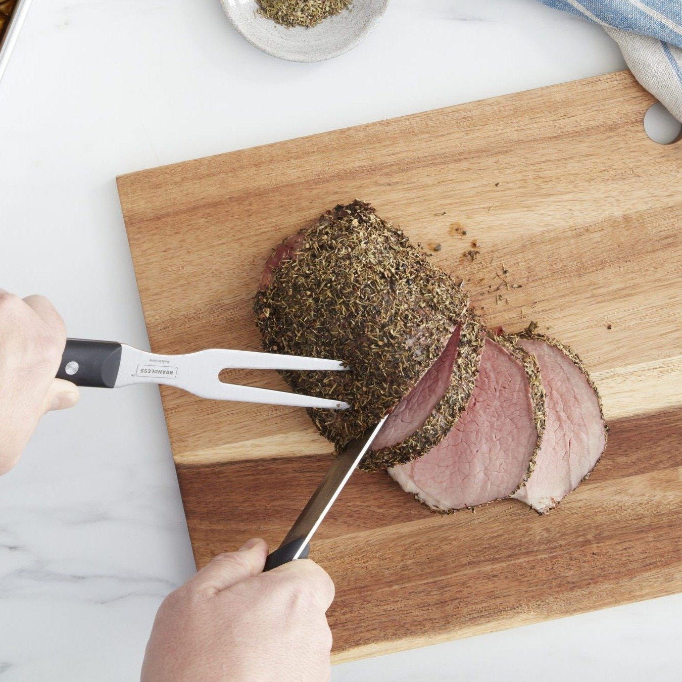 Lifestyle photo, a man's hands using a carving knife and carving fork to neatly slice a pork loin on a cutting board.