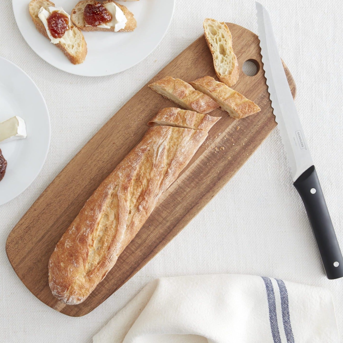 Lifestyle photo, baguette on acacia cheese board, baguette is partially sliced for the tapas with cheese and strawberry compote seen on a nearby serving plate.