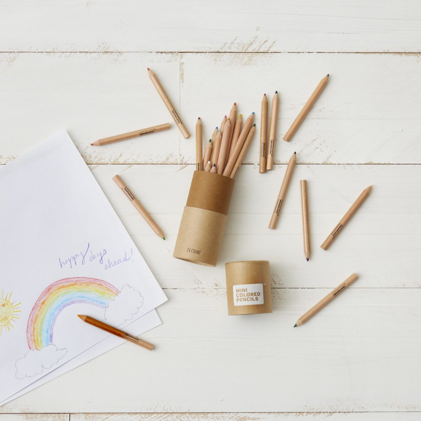 Lifestyle photo, showing a child's drawing of a rainbow next to a canister of mini colored pencils with many colors strewn about during the creative process of drawing.