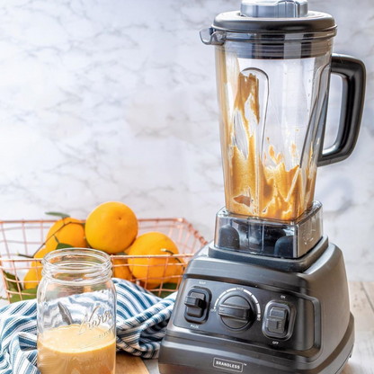 Lifestyle photo, blender on a kitchen countertop next to a basket of oranges, a dishcloth, and a Mason jar with a small batch of freshly-blended peanut butter in it.