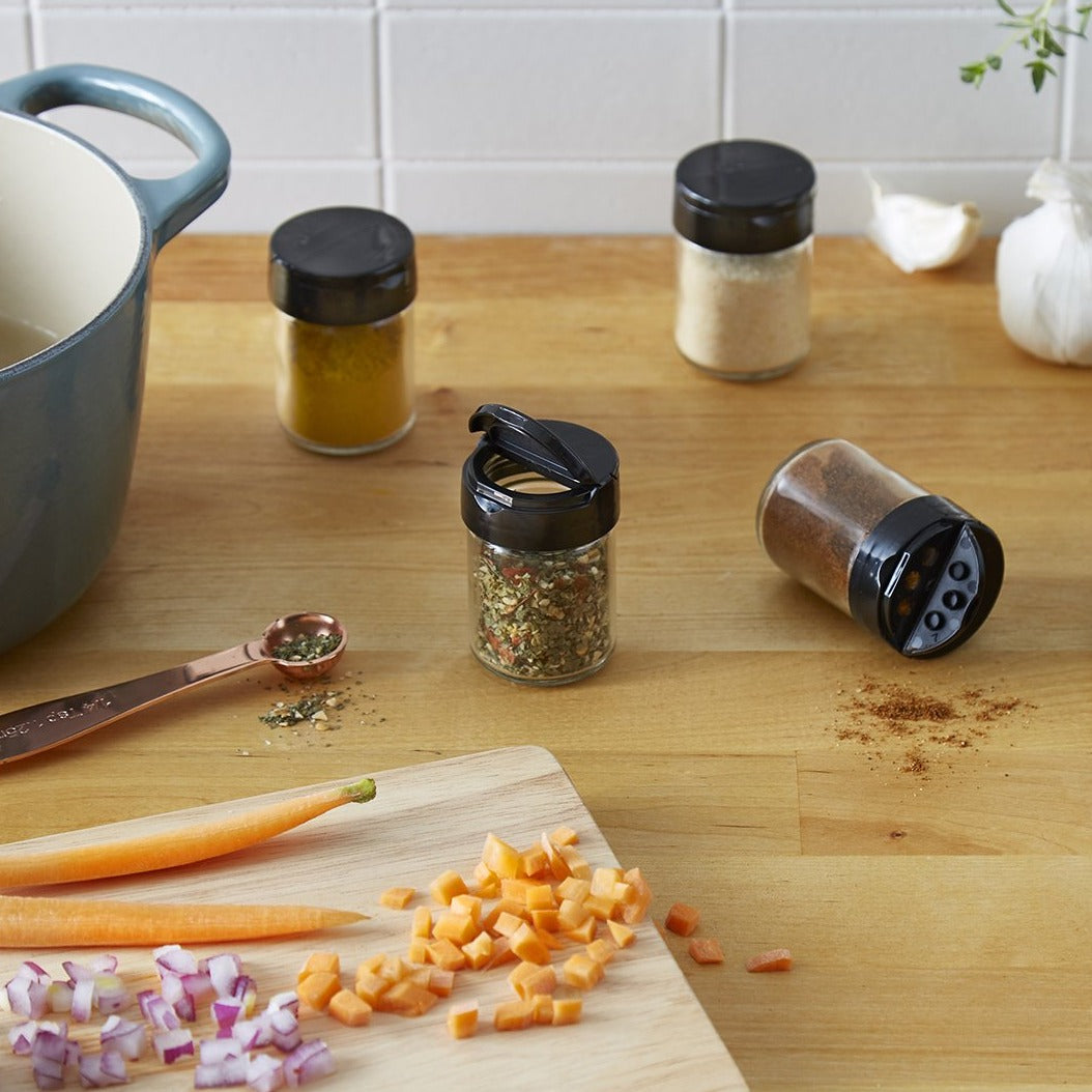 Lifestyle photo. 4 spice jars with various spices in them sit on a kitchen counter amidst the chaos of the kitchen, next to a caserole dish, diced carrots and onions.